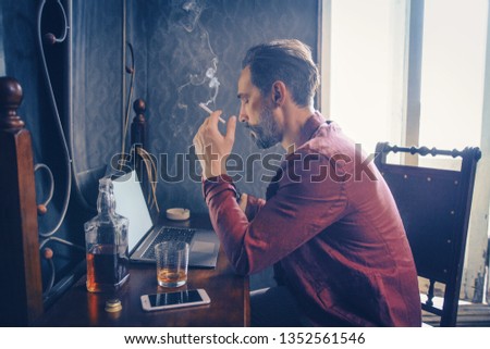 Concentrated Bearded Man Holds A Cigarette While Working On Laptop. Handsome Guy Holds A Cigarette Smoking. Bottle Of Whiskey, Smartphone And Opened Laptop Are On The Wooden Table With A Mirror. Side