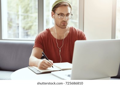 Concentrated Bearded Young European Hipster In Stylish Round Glasses And Green Hat Looking At Laptop Screen And Making Notes In Copybook While Learning Online, Watching Webinar Using Earplugs