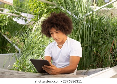 Concentrated beautiful ethnic young man, dressed in casual clothes, works and draws on a tablet outdoors, in a green area in nature. Green open space for student, coworking, freelancer, relaxation - Powered by Shutterstock