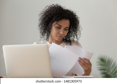 Concentrated biracial millennial girl sit at desk work on laptop reading paperwork education handouts, focused african American young woman busy studying on computer look through paper material - Shutterstock ID 1481305973
