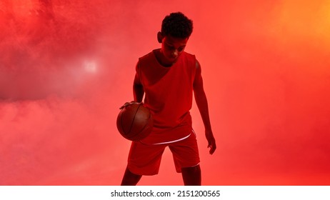 Concentrated Black Boy Basketball Player Dribbling With Ball. Male Youngster Wear Professional Sportswear. Modern Healthy And Sports Lifestyle. Red Background With Smoke. Studio Shoot. Copy Space