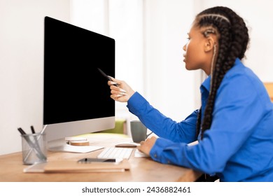 Concentrated black female student with braided hair using blank computer screen, pointing with pen in modern home office setting, doing home assignment - Powered by Shutterstock