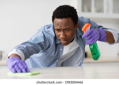 Concentrated Black Guy Cleaning Kitchen Furniture, Using Spray Bottle With Disinfestor And Dust Cloth, Wearing Colorful Rubber Gloves, Copy Space. Young African American Man Cleaning House