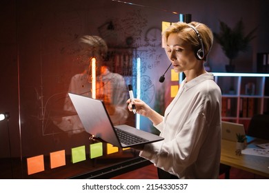 Concentrated Blond Businesslady Writing Tasks And Ideas On Glass Wall With Colorful Stickers At Office. Focused Woman In Headset Using Laptop For Checking Working Progress, Working With Kanban Board