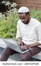 Concentrated Busy Young Muslim Man In Kufi Cap Sitting On Curb And Working With Laptop