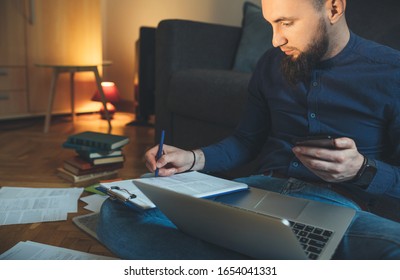 Concentrated Caucasian Businessman With Long Beard Is Writing Something While Using A Laptop And His Phone