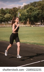 Concentrated Caucasian Girl Athlete Dressed In Sportswear: Shorts And Top, Sneakers With Fitness Tracker Running On Track At City Stadium In Hot Summer. Happy Female Outdoor Workout  Sport Concept