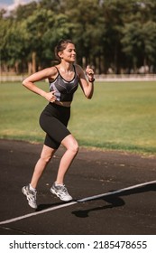 Concentrated Caucasian Girl Athlete Dressed In Sportswear: Shorts And Top, Sneakers With Fitness Tracker Running On Track At City Stadium In Hot Summer. Happy Female Outdoor Workout  Sport Concept