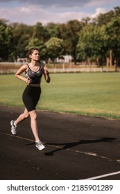 Concentrated Caucasian Girl Athlete Dressed In Sportswear: Shorts And Top, Sneakers With Fitness Tracker Running On Track At City Stadium In Hot Summer. Happy Female Outdoor Workout  Sport Concept