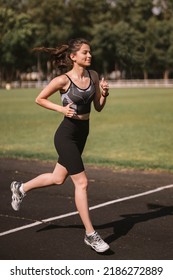 Concentrated Caucasian Girl Athlete Dressed In Sportswear: Shorts And Top, Sneakers With Fitness Tracker Running On Track At City Stadium In Hot Summer. Happy Female Outdoor Workout  Sport Concept