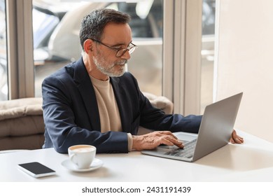 Concentrated caucasian mature businessman with glasses typing on a laptop in a cafe, with a cup of coffee and smartphone on the table, exemplifying modern work flexibility - Powered by Shutterstock