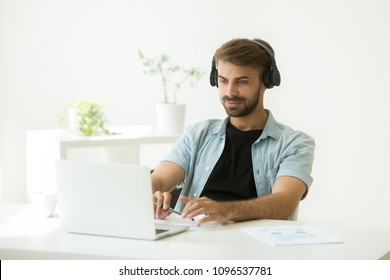 Concentrated Caucasian Worker In Headphones Looking At Laptop Screen, Watching Webinar, Listening To Speaker, Making Notes At Home Or In Office Room. Male Student Taking Online Course, Remote Training