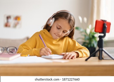 Concentrated child girl doing homework at her room, learning at home, using smartphone with tripod and headphones for remote education with online school teacher or personal tutor. Homeschooling