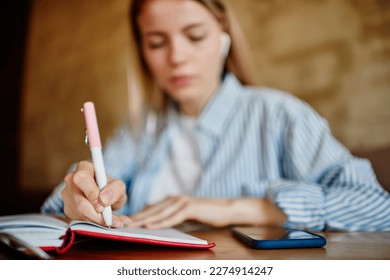 Concentrated crop young female student in casual clothes sitting at table and writing notes in notebook while doing homework and listening to online class on smartphone in cafe - Powered by Shutterstock