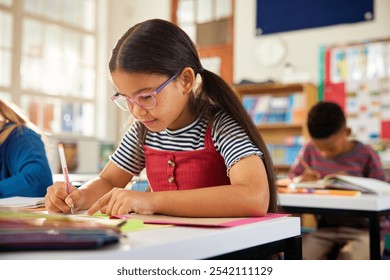 Concentrated cute little primary schoolgirl writing in book with classmates in background at classroom. Little smart pupil writing at desk in classroom at the elementary school. - Powered by Shutterstock