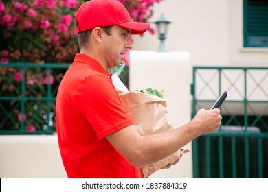 Concentrated deliveryman walking and reading address on tablet. Side view of courier delivering order in paper bag and wearing red shirt and cap. Delivery service and online shopping concept