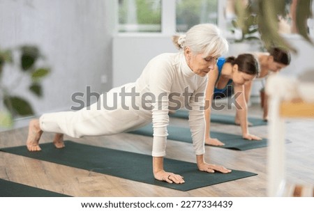 Concentrated elderly woman holding plank pose to strengthen body muscles during group yoga training in studio. Core exercises for older adults. Active lifestyle concept..