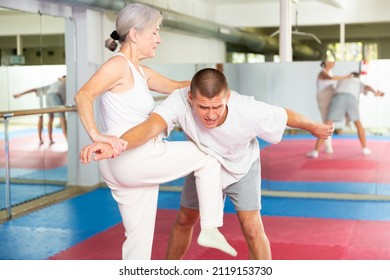 Concentrated elderly woman practicing self defense techniques, performing elbow and knee strikes with arm hold to male sparring partner in gym