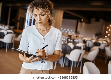 concentrated event organizer writing checklist on clipboard near festive tables in banquet hall