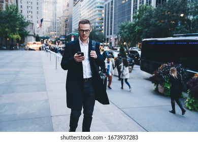 Concentrated Executive In Formal Wear And Trendy Glasses Using Smartphone While Walking Down Crowded Street And Carrying Bag On Shoulder