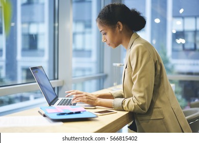 Concentrated female administrative typing messages to employee coordinating work being in business trip using laptop computer and wireless connection to internet sitting in modern coworking space