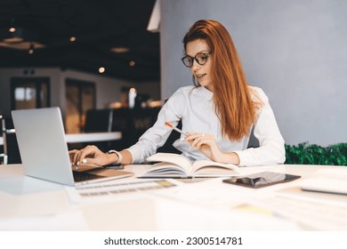 Concentrated female employee in smart casuals and eyeglasses sitting at table with diary and using laptop while working on project in office - Powered by Shutterstock