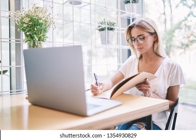 Concentrated female journalist in eyeglasses for vision correction writing questions for next interview using textbook for education, young student learning online with webinar on laptop computer