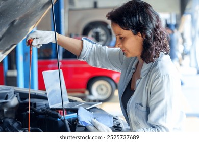 Concentrated female mechanic checking oil dipstick in car engine while standing in an automotive workshop, wearing gloves and casual clothing, manual car maintenance skills and professional expertise. - Powered by Shutterstock