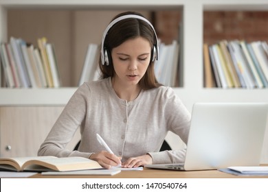 Concentrated female student sitting at desk wearing wireless headphones, enjoying favorite music tracks during study or listening online lecture, writing down notes, preparing for seminar or exam. - Shutterstock ID 1470540764