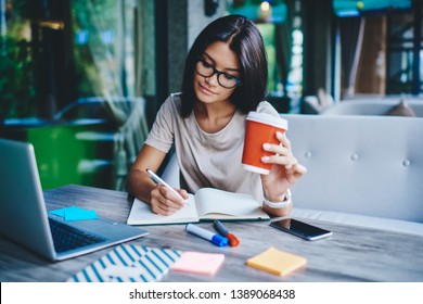 Concentrated female student writing in notebook while learning with cardboard coffee cup in cafe, pensive woman freelancer noting information for planning project doing remote job via laptop computer