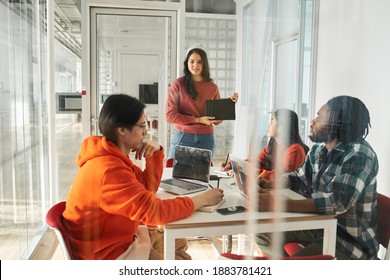 Concentrated Female Team Leader Standing In Front Of The Table And Showing Something On The Tablet To Explain New School Project Details To Diverse Students. Group Of Mixed Race Happy Young People