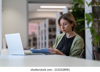 Concentrated Female University Professor Sitting In Front Of Laptop In Empty Library Writing Down Notes While Preparing For Lecture Or Checking Course Schedule, Selective Focus. Academic Career