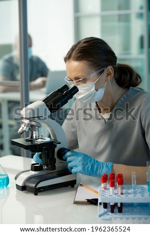 Concentrated female virologist in protective goggles and latex gloves sitting at desk in laboratory and learning cells under microscope