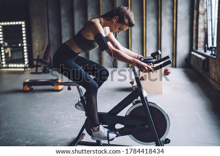 Concentrated fit female in sportswear with dark braided hair burning calories on spin bike and listening to music in headphones
