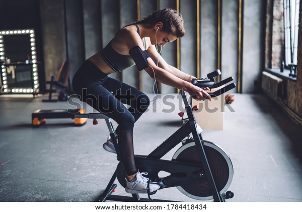Concentrated fit female in sportswear with\
dark braided hair burning calories on spin bike and listening to\
music in\
headphones