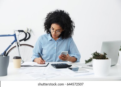 Concentrated focused Afro American female freelancer holding phone in one hand and making notes with pen in other while planning budget and calculating bills, sitting at desk with papers and gadgets
