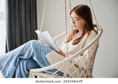 Concentrated girl relaxing and reading book on indoor swing. Pretty female in glasses excited about story she reading. Cozy afternoon with book in hanging chair in home. - Powered by Shutterstock