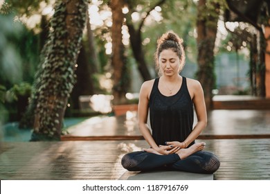 Concentrated girl sitting in lotus pose and meditating or praying. Young woman practicing yoga alone on wooden deck in tropical island - Powered by Shutterstock