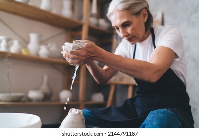 Concentrated grey hair caucasian female sculptor making clay pot on pottery wheel. Concept of small business and entrepreneurship. Home hobby, entertainment and leisure. Woman working at art studio - Powered by Shutterstock