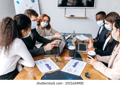 Concentrated Group Of Business People At Briefing Meeting. Multiracial Coworkers Wearing Medical Masks, Studying At Financial Graphs, Brainstorming Together, Discuss Ideas, Working On New Project