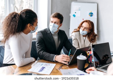 Concentrated Group Of Business People At Briefing Meeting. Multiracial Coworkers Wearing Medical Masks, Studying At Financial Graphs, Brainstorming Together, Discuss Ideas, Working On New Project