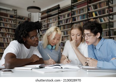 Concentrated group of multiethnic young students reading textbook, writing notes preparing for seminar or exams together, improving knowledge sitting at table in modern library or classroom.
