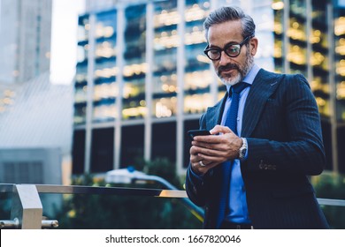 Concentrated handsome bearded businessman with trendy haircut in black coat and blue tie in spectacles texting on smartphone by glass fence on blurred background in New York 