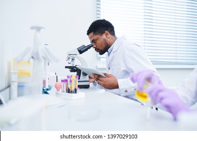 Concentrated Handsome Black Male Lab Technician With Beard Sitting At Table And Using Microscope While Analyzing Sample And Working With Data On Tablet