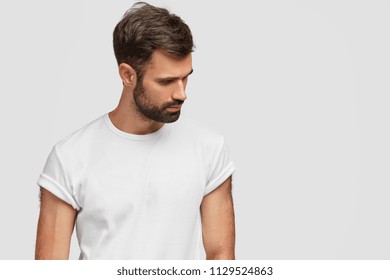Concentrated handsome young male focused down, has dark stubble and hair, wears casual white t shirt, isolated over studio background with copy space for your advetisement or promotional text