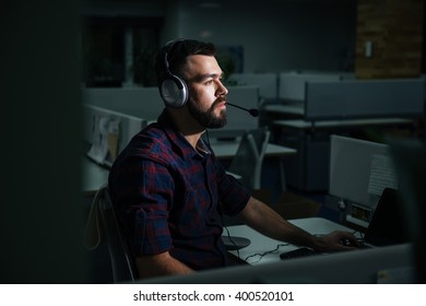 Concentrated Handsome Young Man In Headphones Sitting And Working At Night In Dark Office 