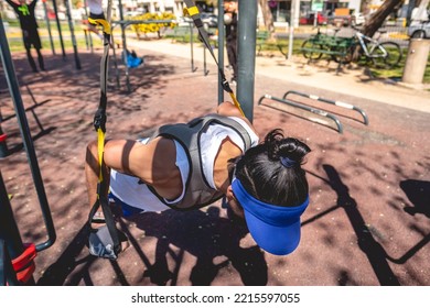 Concentrated, Healthy And Fit Young Latin Man With Weight Vest, Workout Gloves And Blue Shorts And Visor Performing Exercises With Elastic Bands On Bars In A Workout Park In Sunny Day