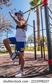 Concentrated, Healthy And Fit Young Latin Man With Weight Vest, Workout Gloves And Blue Shorts And Visor Performing Exercises With Elastic Bands On Bars In A Workout Park In Sunny Day