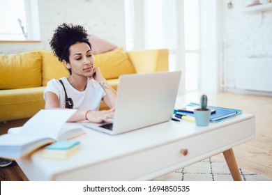 Concentrated Hispanic Female In Casual Wear Browsing On Internet Using Laptop While Sitting On Floor In Light Filled Room At Home