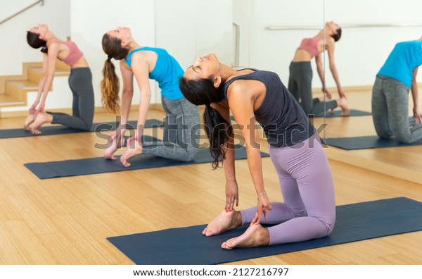 Concentrated hispanic woman performing kneeling\
back-bending asana Ustrasana (Camel Pose) during group yoga course\
in fitness studio